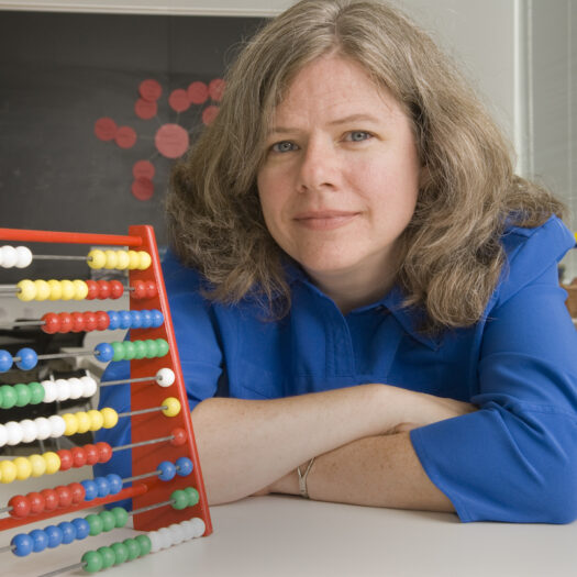 Jennifer McCray smiles and poses for her headshot. She is seated next to an abacus and is wearing a bright blue blouse.